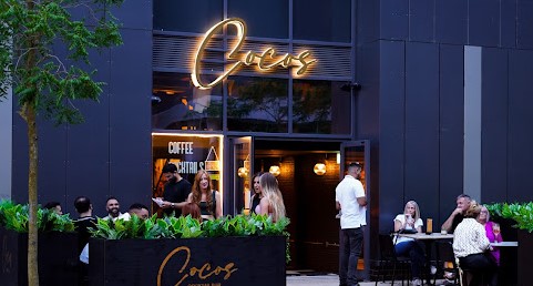 Cocos exterior with dark grey walls with gold LED sign, outdoor seating surrounded by plants