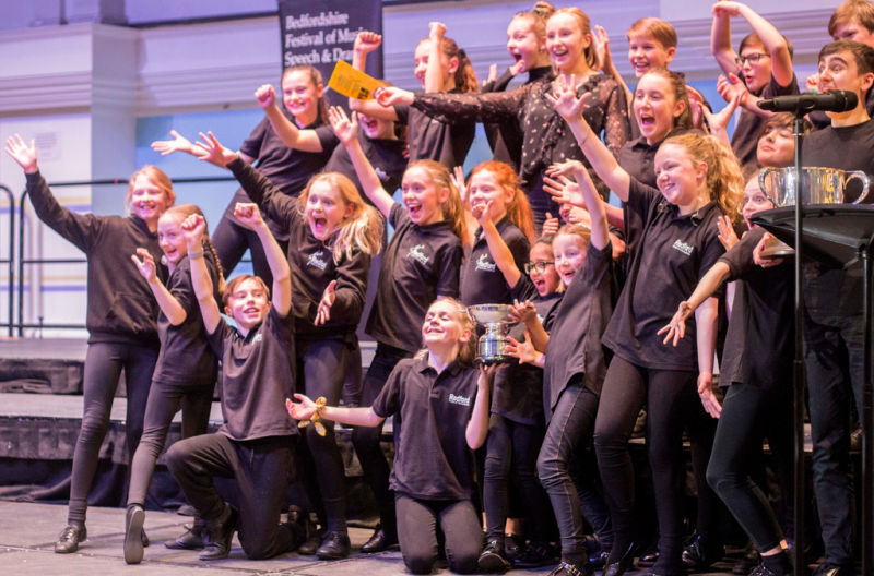 Bedfordshire Festival of Music Speech and Drama children on a stage performing with theor arms raised.