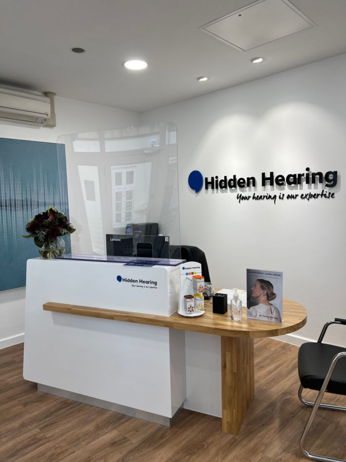 White reception counter with wooden desk. White walls with mounted Hidden Hearing signage behind.