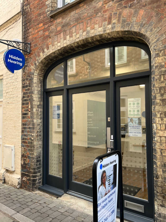 Exterior entrance with display board. Black wooden framed glass windows and door surrounded by brickwork